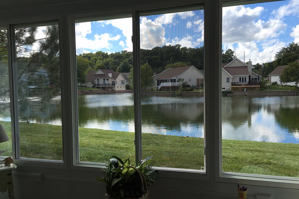 View through large windows showing a grassy area, a pond, and houses on the opposite shore under a partly cloudy sky. Inside, a plant in a basket and part of a lamp and table are visible.