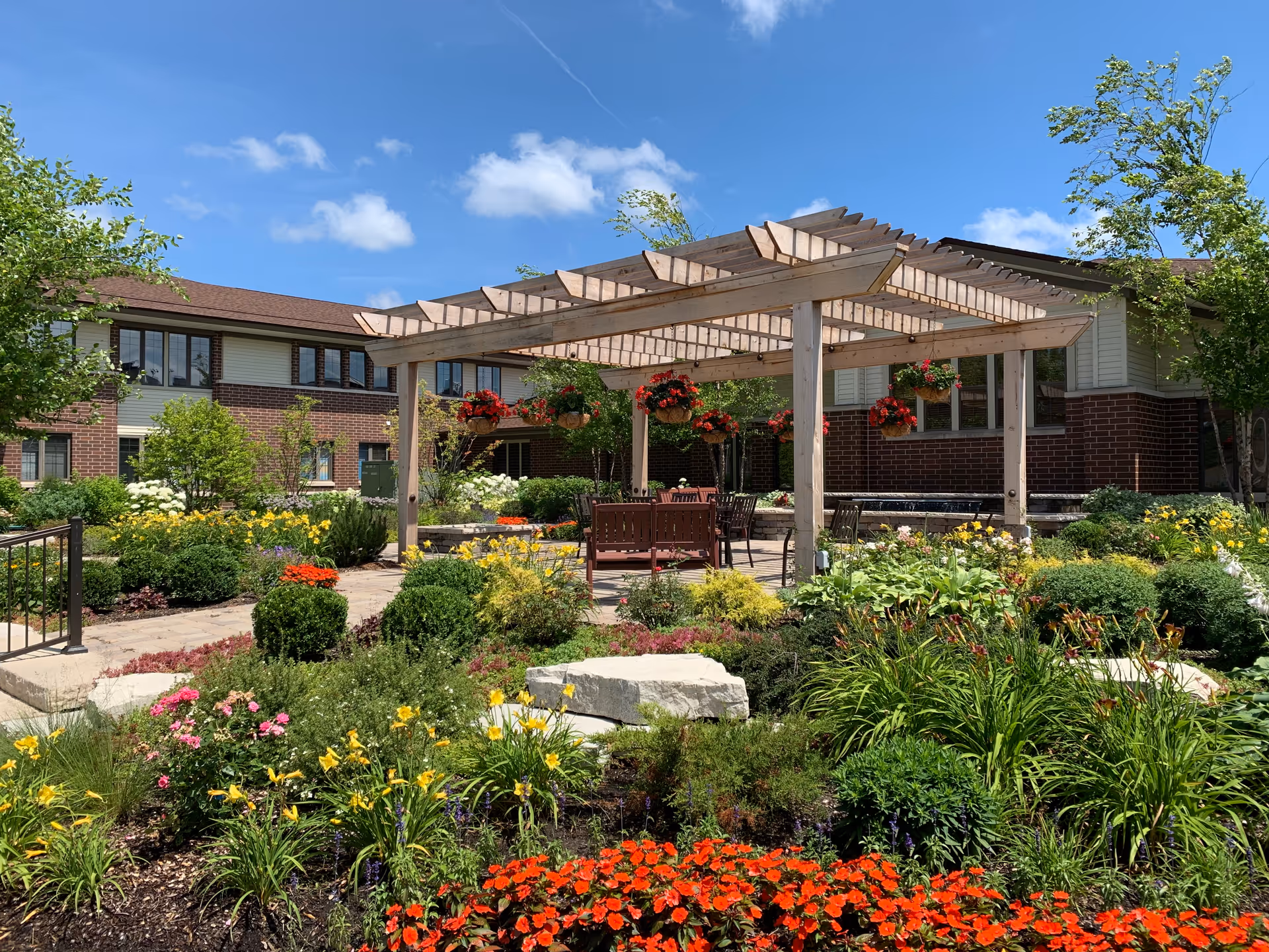 Outdoor garden area at Village At Victory Lakes with a wooden pergola, hanging flower baskets, various colorful flowers, shrubs, and patio furniture under a clear blue sky.