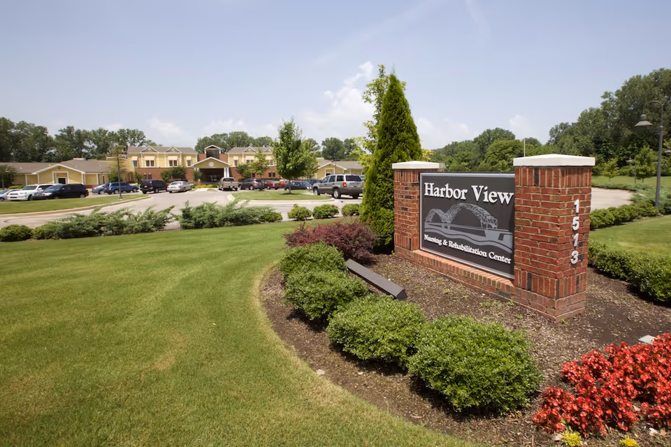 Exterior view of AHC Harbor View Nursing & Rehabilitation Center with a brick sign in the foreground surrounded by landscaped bushes and flowers, and the facility building and parking lot with cars in the background under a partly cloudy sky.