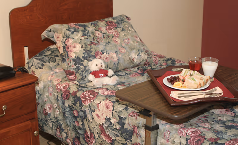 A hospital-style bed with floral patterned bedding and a small white teddy bear wearing a red shirt sitting on the bed. A wooden bedside table with a black telephone is next to the bed. A wooden overbed table holds a tray with a plate of sandwiches, grapes, an orange slice, a glass of red juice, and a glass of milk.