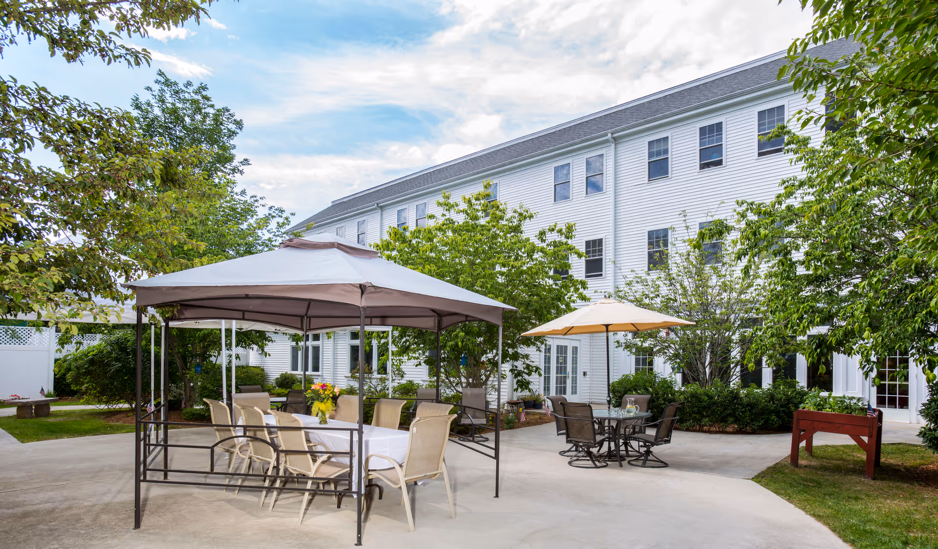 Outdoor patio area at Benchmark Senior Living at Robbins Brook featuring a large gazebo with a table covered by a white tablecloth and surrounded by chairs, another smaller table with an umbrella and chairs, lush green trees and bushes, and a white multi-story building in the background under a partly cloudy sky.