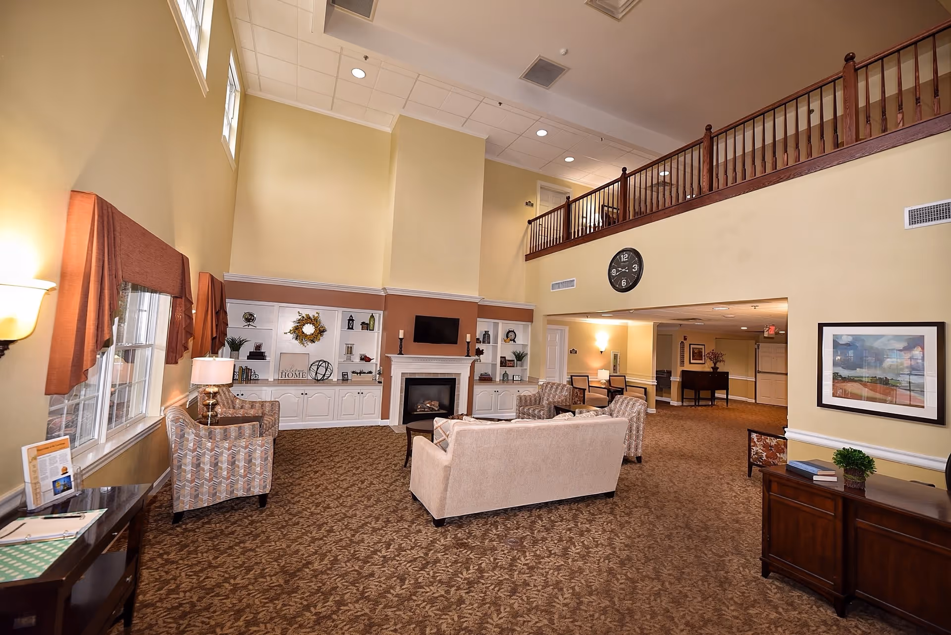 A spacious senior living facility common area with high ceilings, beige walls, and patterned carpet. The room features a beige sofa, two patterned armchairs, a fireplace with a mounted TV above it, built-in white shelves decorated with plants and ornaments, and a wooden railing on the upper level. There are windows with brown valances on the left side and a clock on the wall above an open hallway.