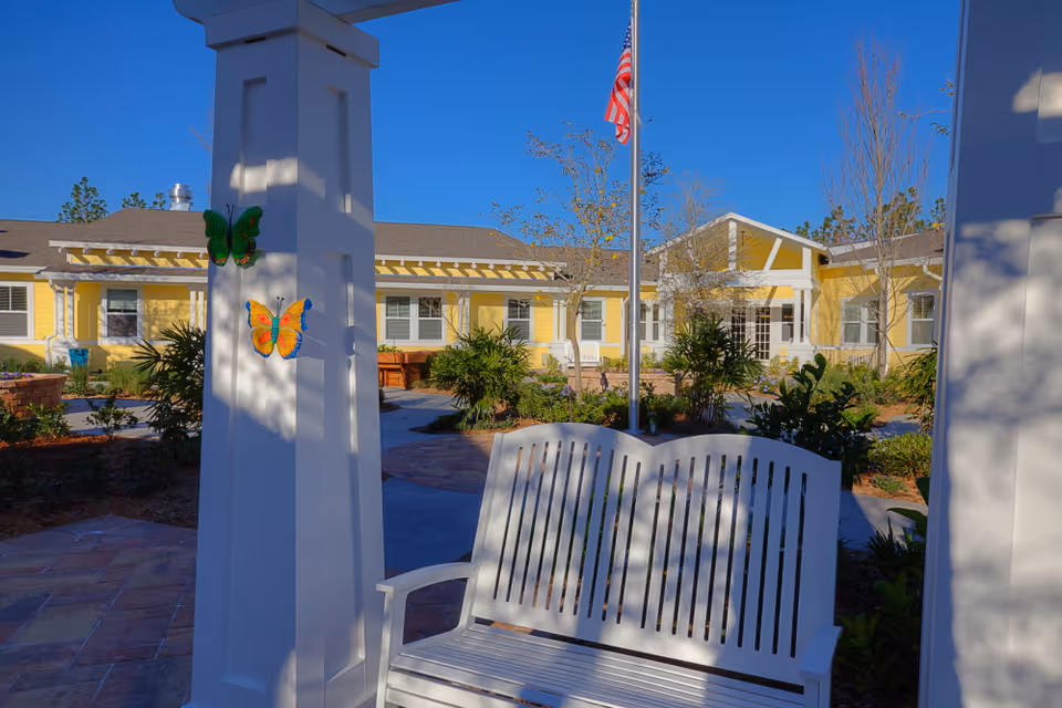 Outdoor courtyard area of a senior living facility with a white bench in the foreground, a white pillar decorated with colorful butterfly ornaments, and a yellow building with white trim in the background under a clear blue sky. An American flag is flying on a flagpole near the center.