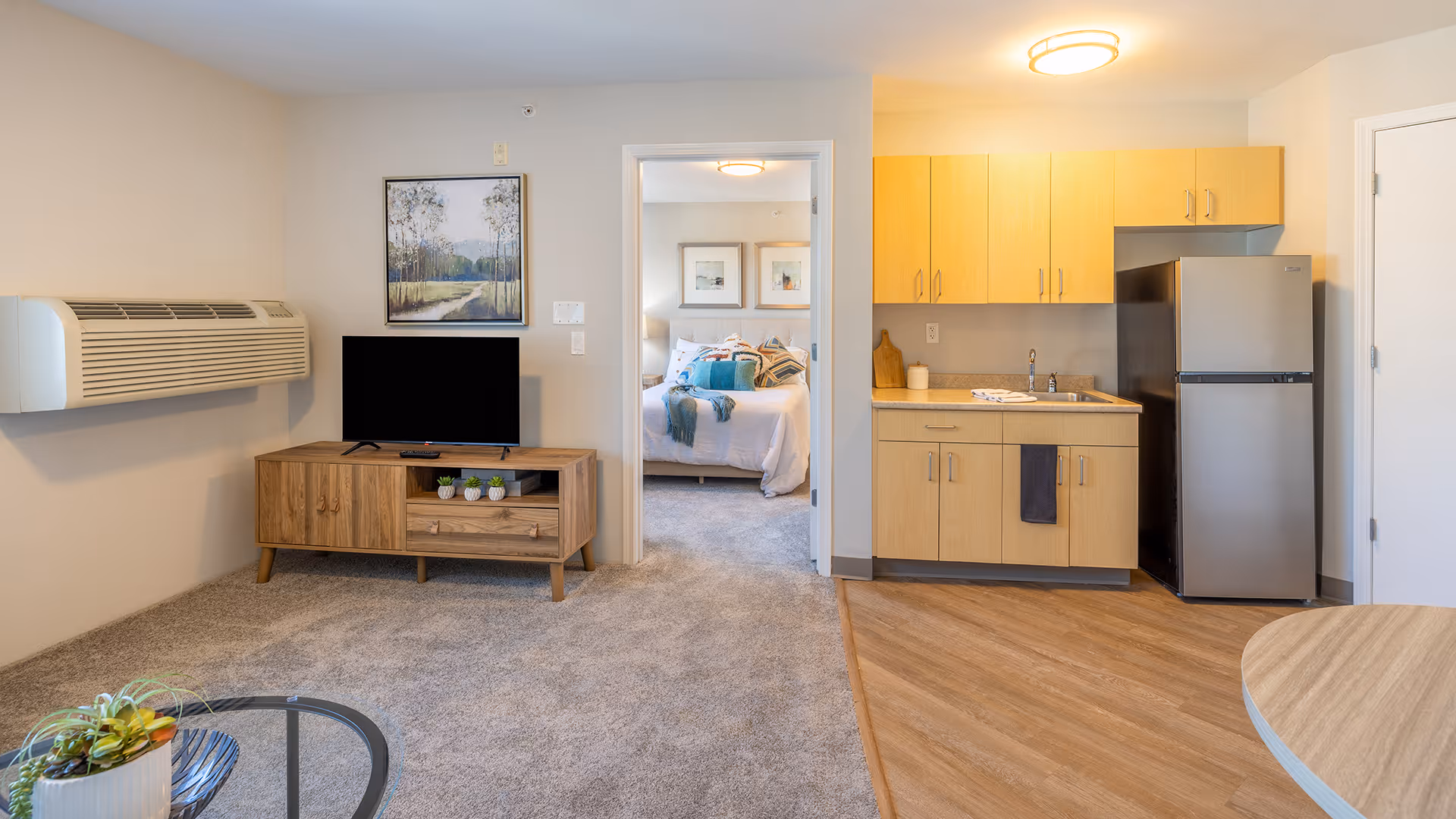 A bright and cozy senior living apartment featuring a small kitchenette with light wood cabinets, a stainless steel refrigerator, and a sink. Adjacent to the kitchenette is a living area with a wooden TV stand holding a flat-screen television and decorative plants. The room has beige walls, carpeted flooring in the living area, and wood flooring in the kitchenette. A doorway leads to a bedroom with a neatly made bed adorned with pillows and blankets, and framed artwork on the walls.