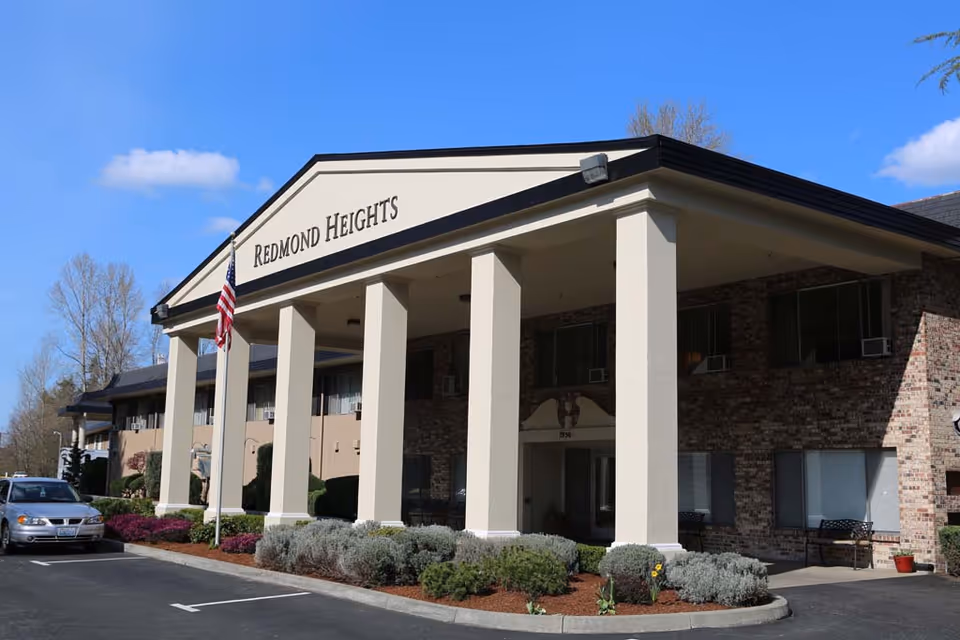 Exterior view of Redmond Heights Senior Living building with a covered entrance supported by large white columns, an American flag on a flagpole, and a parked car in front. The building has brick walls and several windows with air conditioning units.
