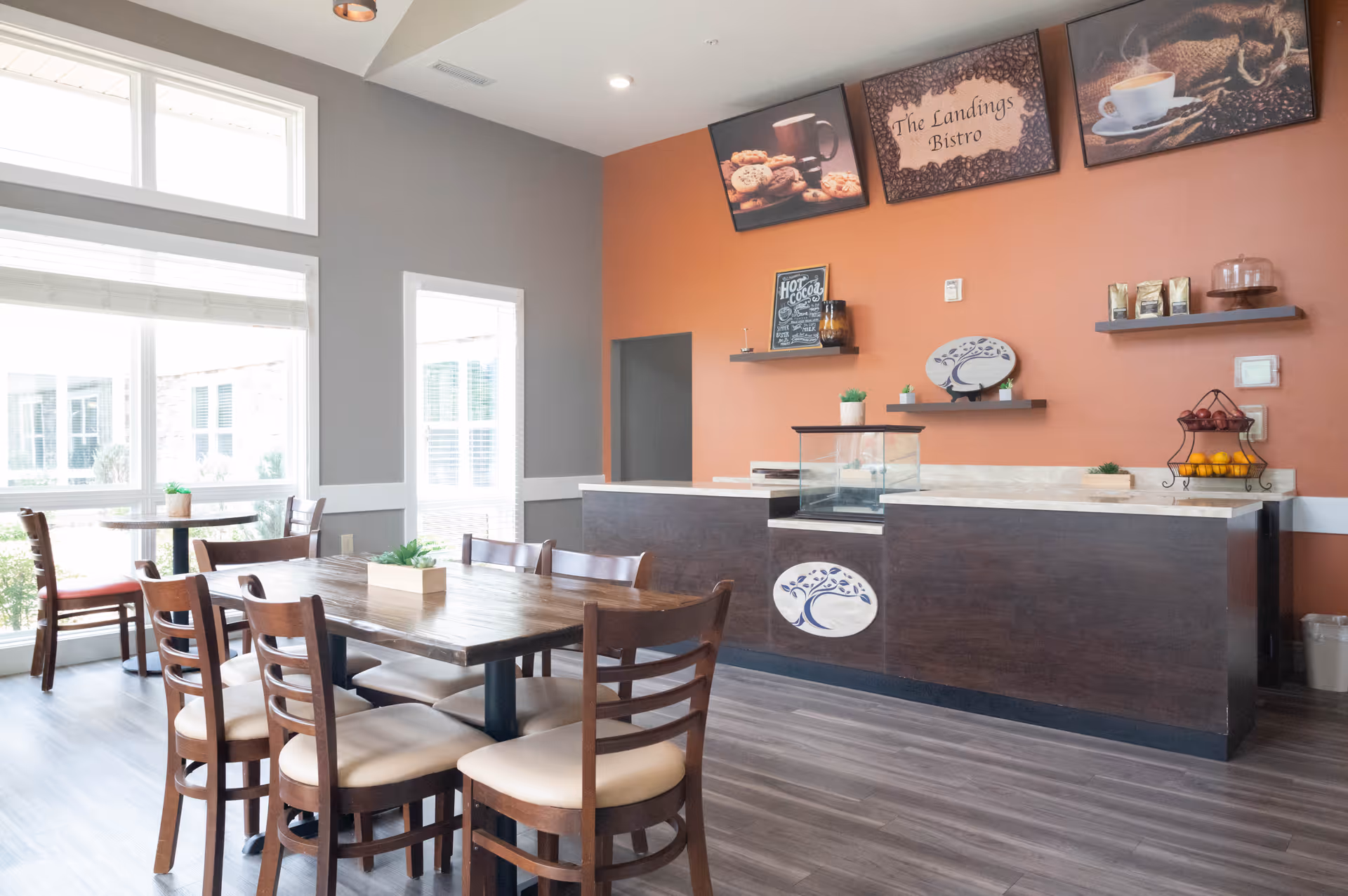 A bright dining area with wooden tables and chairs, large windows letting in natural light, and a counter labeled 'The Landings Bistro' with decorative shelves and framed pictures of coffee and cookies on the orange accent wall.