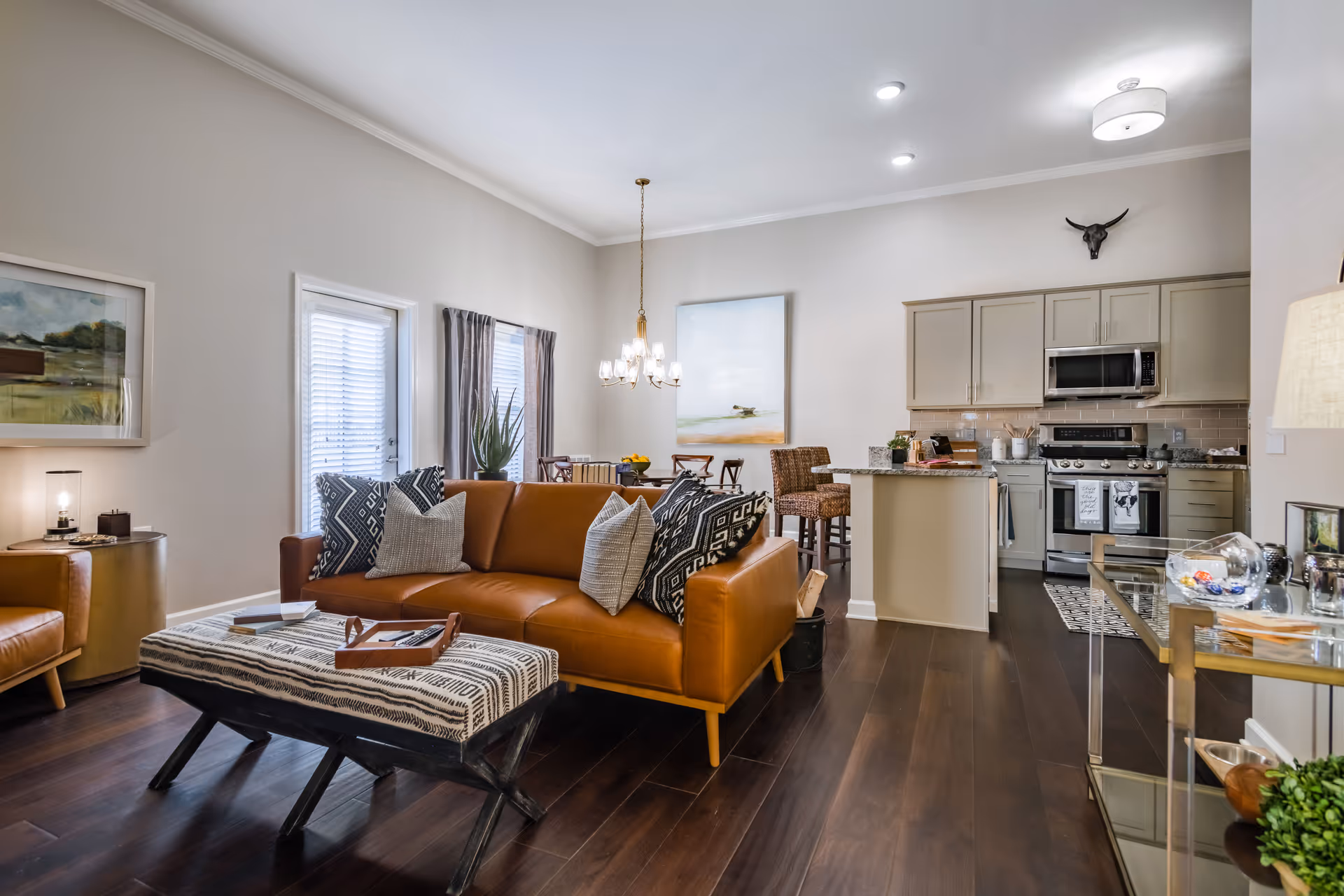 Open-concept living area with a brown leather sofa and patterned ottoman facing a dining table and a kitchen with an island.
