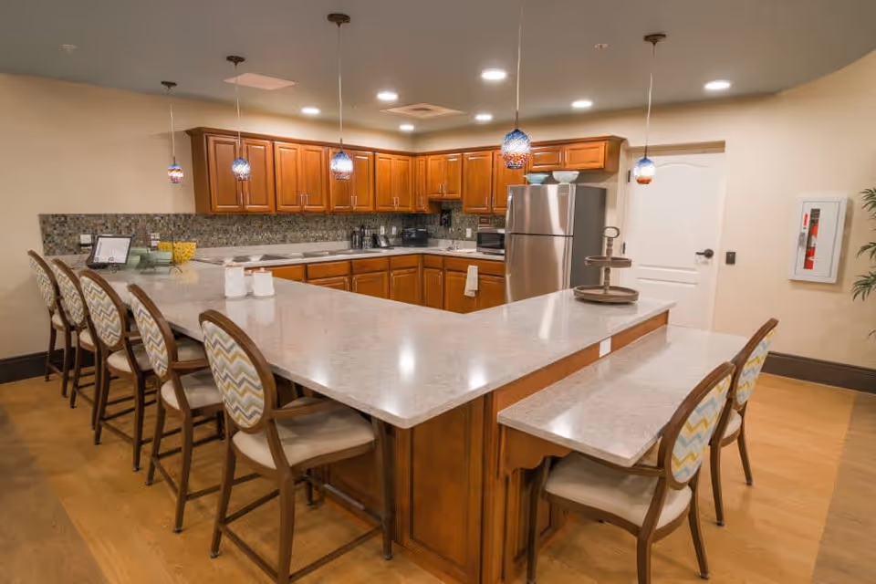 A spacious kitchen area with a large U-shaped countertop surrounded by eight chairs with patterned upholstery. The kitchen features wooden cabinets, a stainless steel refrigerator, a microwave, and a tiled backsplash. Five pendant lights hang from the ceiling above the counter, and the floor is wooden.