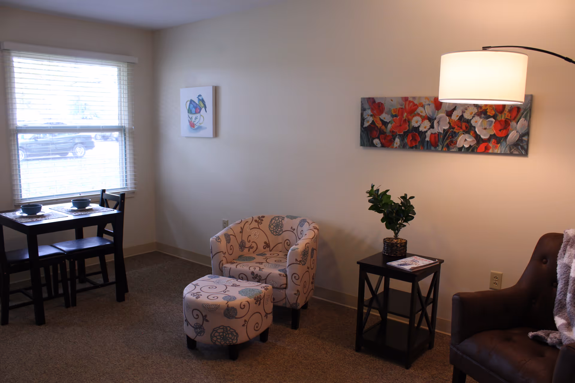 Bright living room with a patterned armchair and ottoman, small side table with a plant, a dining table by a window, and floral wall art.