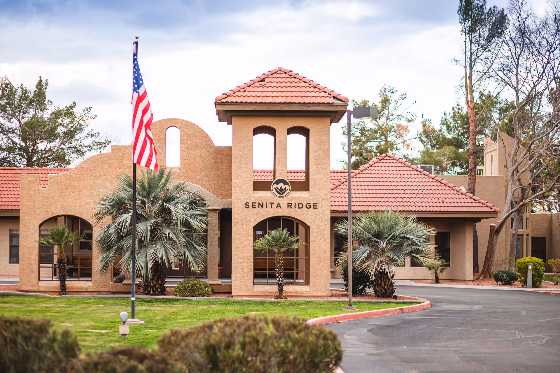 Exterior view of Senita Ridge facility with tan stucco walls and red tile roof. The building features arched windows and a central tower with the Senita Ridge logo and name. There are palm trees and other landscaping in front, an American flag on a flagpole, and a paved driveway curving in front of the entrance.
