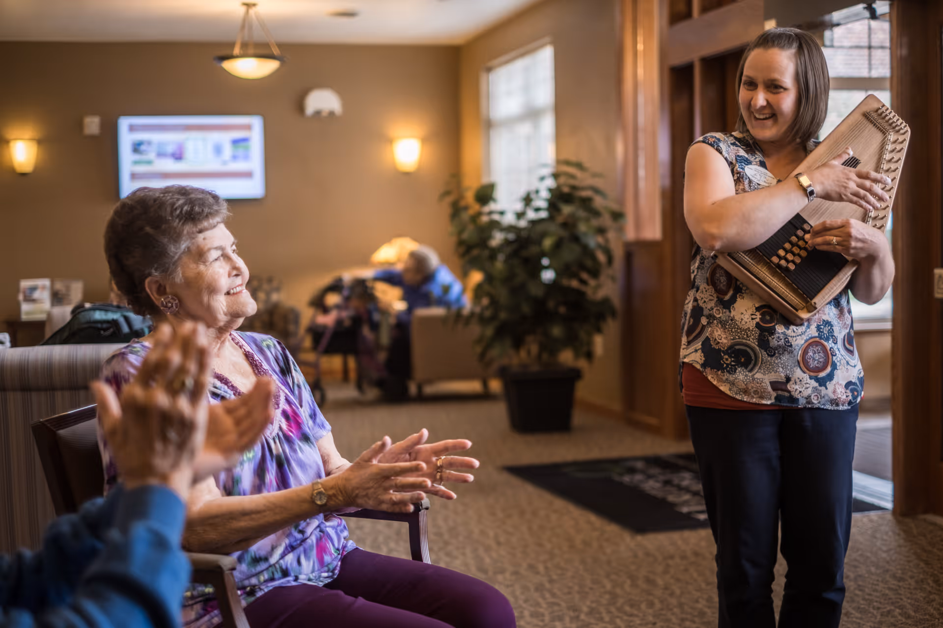 An elderly woman sitting and clapping her hands while smiling, facing a woman standing and holding a musical instrument in a warmly lit common area with other people and plants in the background.