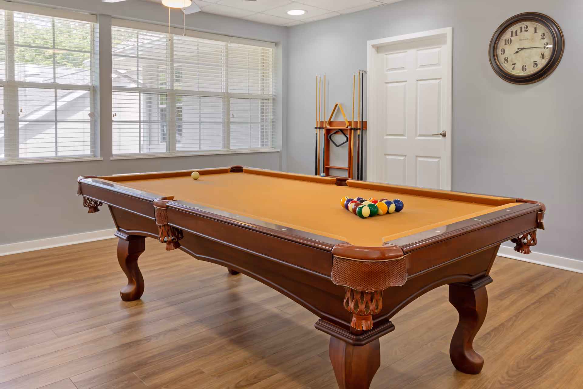 A well-lit room with a wooden pool table featuring a tan felt surface. The pool balls are arranged in a triangle on the table, and several pool cues are stored in a rack against the wall. The room has large windows with white blinds, a white door, a wall clock, and wood flooring.