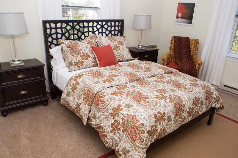 A neatly made bed with a dark wooden headboard and floral patterned bedding in shades of red, beige, and green. There are two matching nightstands on either side of the bed, each with a lamp. To the right of the bed is a wicker armchair with a red cushion and a brown throw blanket. The room has beige carpet and light-colored walls with a framed picture hanging on the wall.