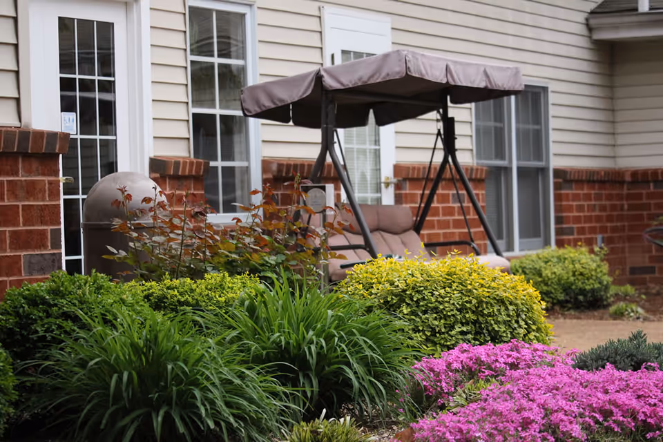 A cushioned patio swing on a landscaped courtyard with shrubs and pink flowers in front of a brick-and-siding building.