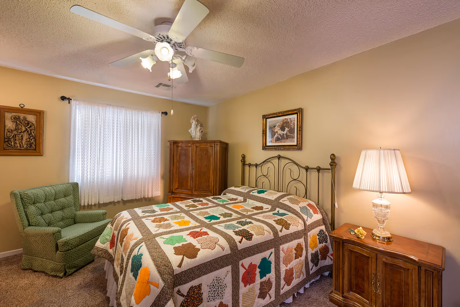 A cozy bedroom with a bed covered in a quilt featuring colorful leaf patterns. There is a green upholstered armchair near a window with white lace curtains. A wooden cabinet stands in the corner, and a wooden nightstand with a decorative lamp and a small flower is beside the bed. The walls are painted beige and decorated with framed artwork. A ceiling fan with lights is mounted on the ceiling.