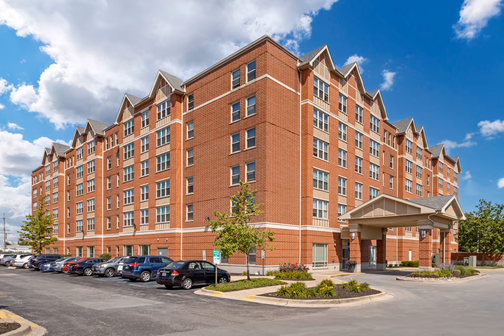 Exterior view of a large multi-story brick building with multiple windows and a covered entrance. Several cars are parked in the parking lot in front of the building under a partly cloudy blue sky.