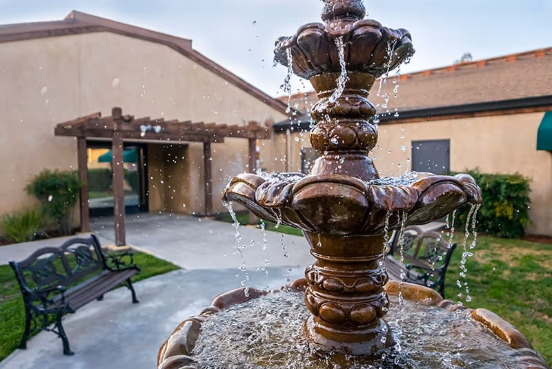 Outdoor courtyard area with a multi-tiered water fountain in the foreground, two black metal benches on either side, a concrete pathway, and a building with beige walls and a wooden pergola in the background.