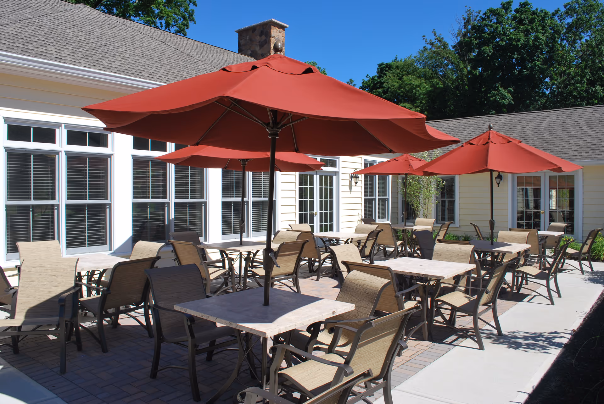 Outdoor patio area with multiple tables and chairs under large red umbrellas next to a building with white-framed windows and beige siding, surrounded by trees and greenery under a clear blue sky.