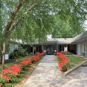 Pathway lined with red flowers leading to the entrance of a single-story building surrounded by green trees and grass under a clear sky.