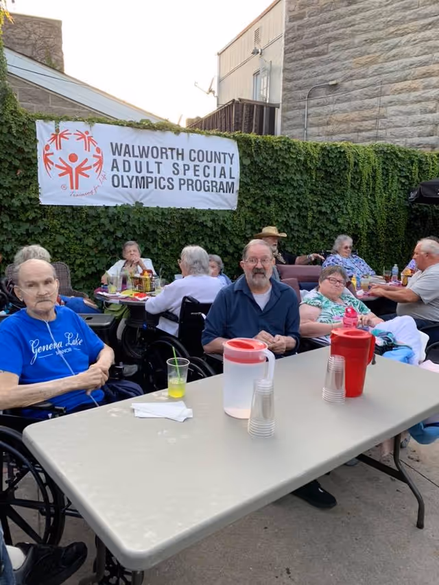 A group of elderly people sitting outdoors at tables with drinks and snacks. Some are in wheelchairs. Behind them is a green ivy-covered wall with a banner that reads 'Walworth County Adult Special Olympics Program'. The setting appears to be a social or community event at Geneva Lake Manor.