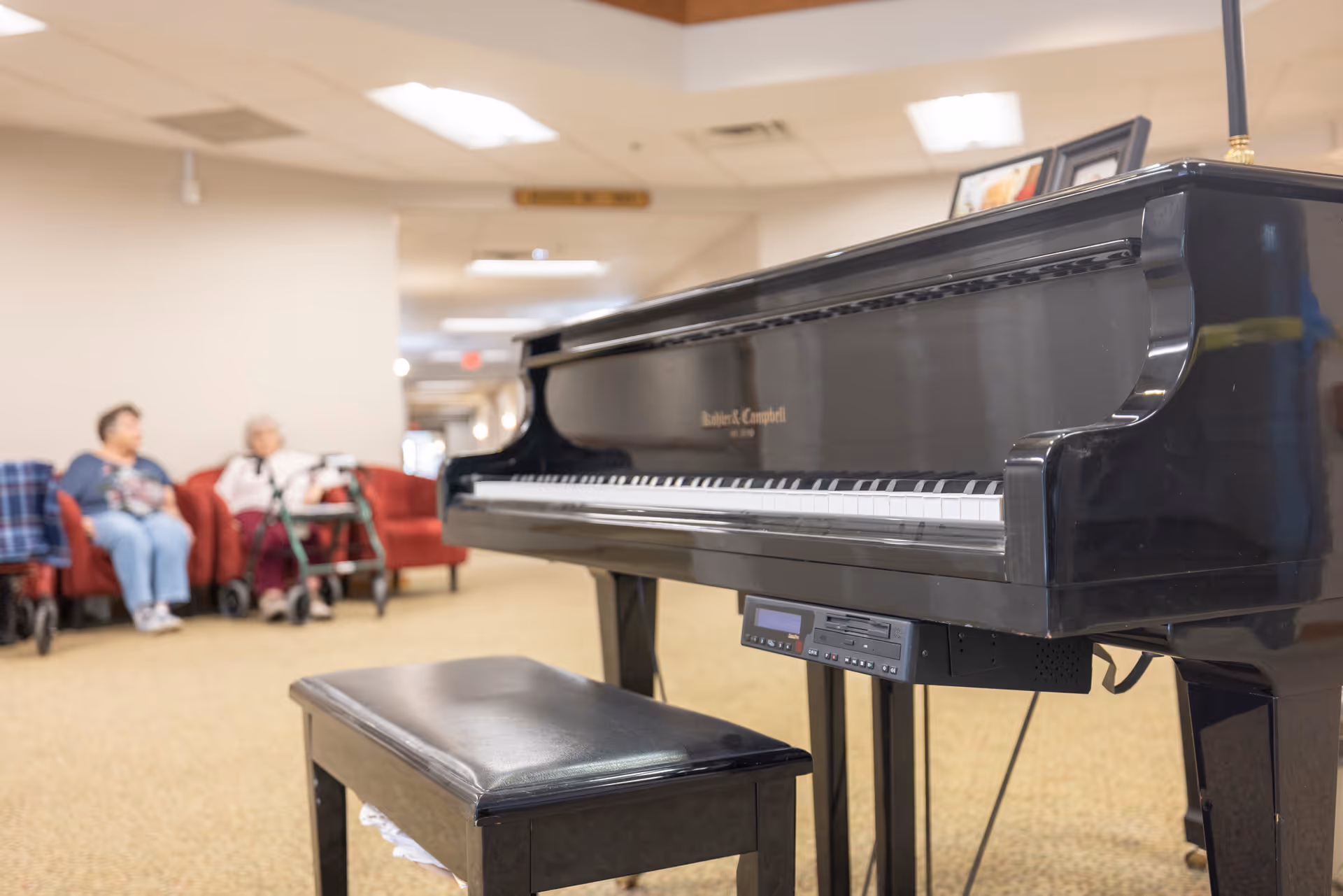 A black grand piano with a matching bench is positioned in a common area of a senior living facility. In the background, two elderly women are seated on red chairs, one with a walker nearby. The room has beige carpet and neutral-colored walls with ceiling lights.