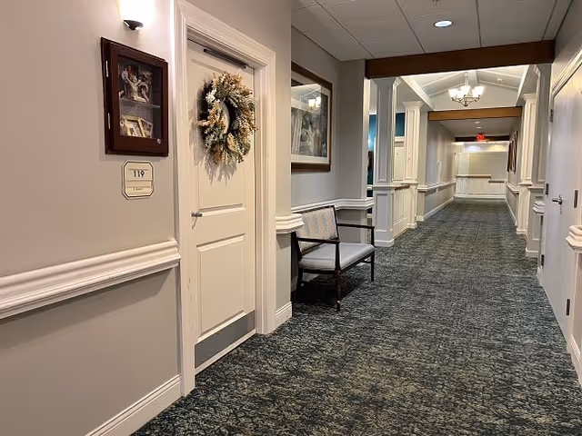 A long, carpeted hallway in a senior living facility with white walls and decorative molding. There is a door on the left side adorned with a wreath and a room number plaque labeled 119. A cushioned bench is placed against the wall beneath a framed picture. The hallway is well-lit with ceiling lights and a chandelier further down the corridor.
