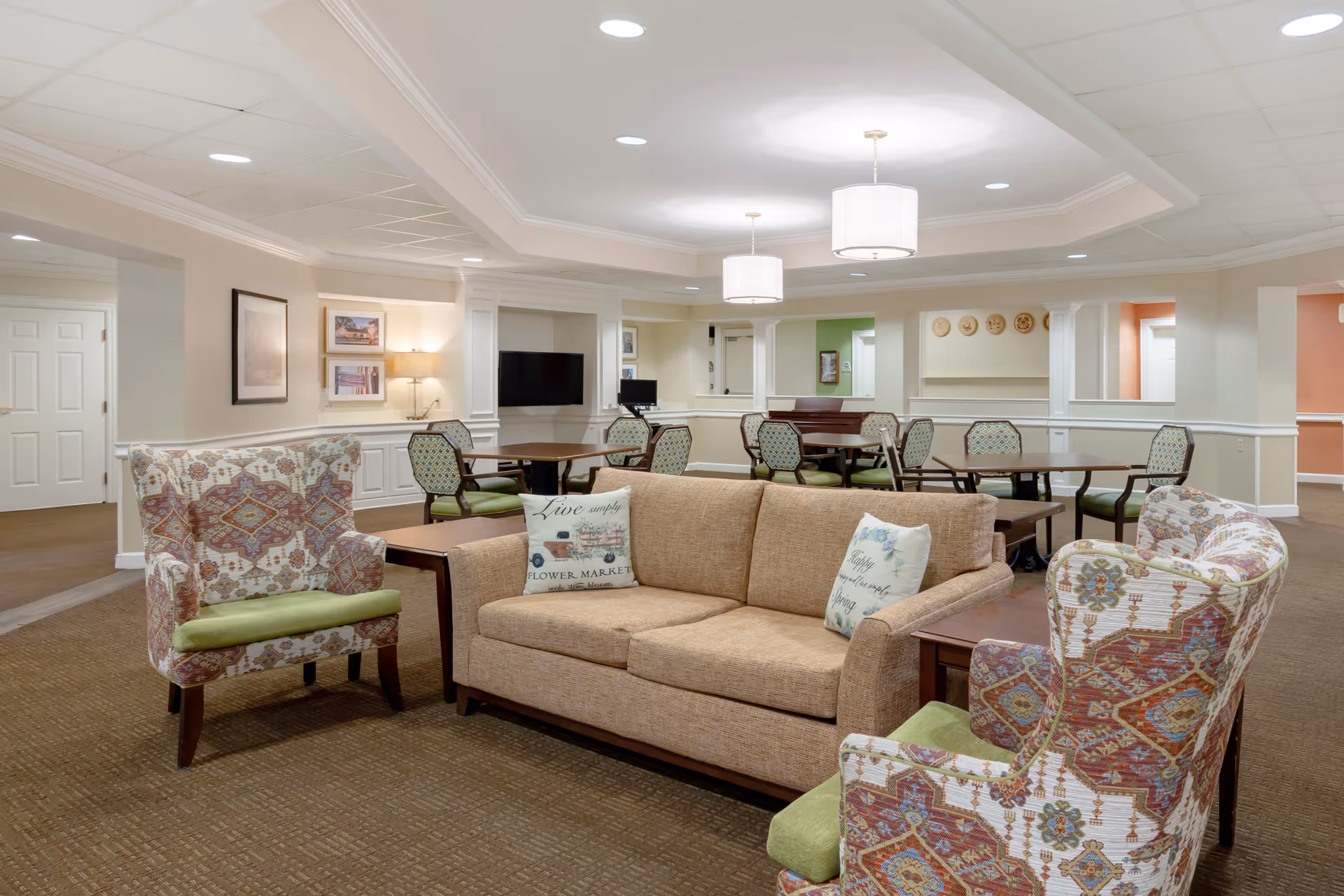 A cozy senior living facility common area with a beige sofa and two patterned armchairs with green cushions arranged around a wooden coffee table. In the background, there are several round tables with chairs, a wall-mounted TV, framed pictures, and soft lighting fixtures on the ceiling and walls.