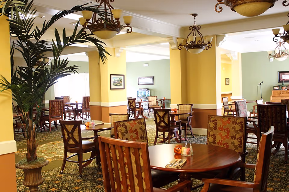 A bright and spacious dining room in a senior living facility with multiple wooden tables and chairs arranged neatly. The room features yellow and green walls, patterned carpet, decorative chandeliers, and a large potted plant. There are framed pictures on the walls and a piano in the background.