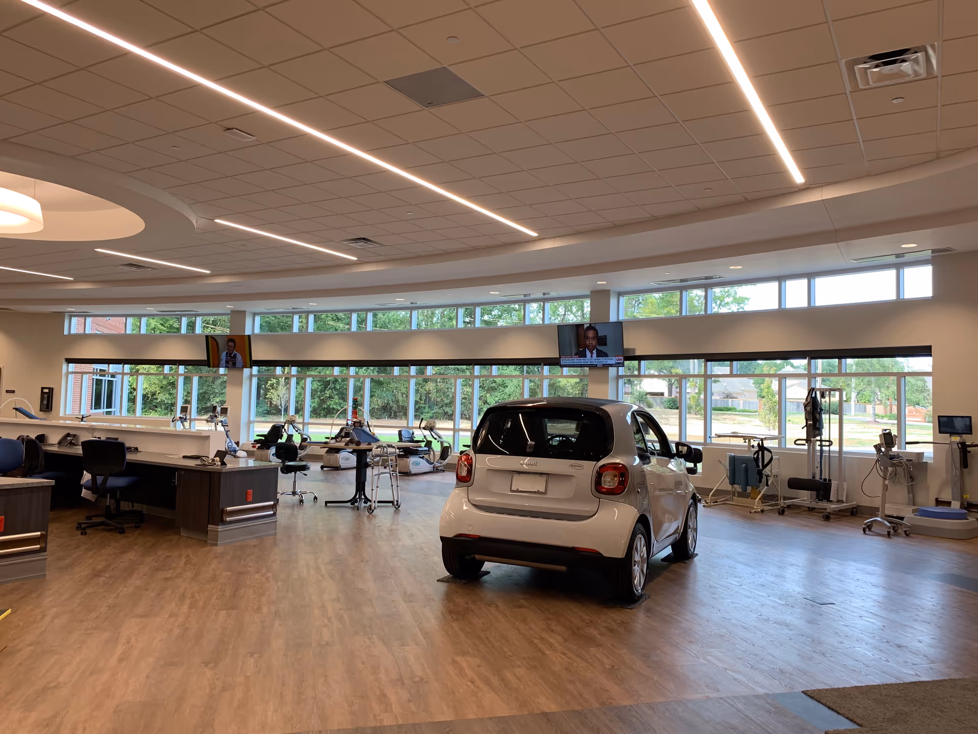 Spacious interior rehabilitation therapy room with exercise equipment, workstations, and a small white car near large windows.
