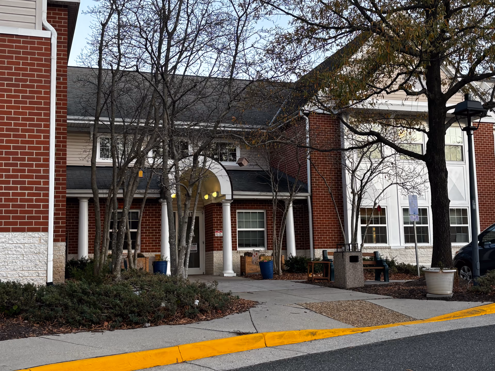 Exterior view of Stevenson Place showing the entrance with white columns and a covered porch. The building has red brick and white siding with several windows. There are leafless trees and some shrubs near the entrance, a bench, a trash can, a street lamp, and a parking area with a car visible on the right.