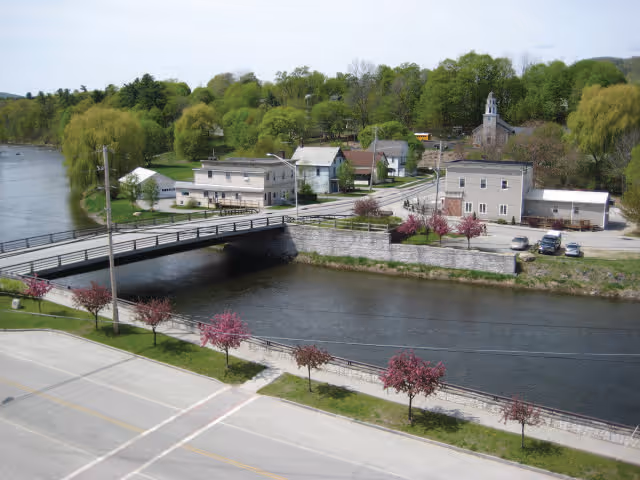 A scenic view of a small town area featuring a river with a stone embankment, a bridge crossing the river, several buildings including houses and a church with a steeple, and a road lined with small trees with pink blossoms.