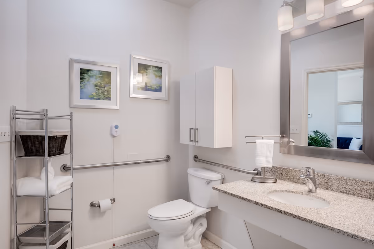 A clean and modern bathroom with a white toilet, a granite countertop with an under-mount sink, a large mirror with three light fixtures above it, a white wall cabinet, a metal towel rack with white towels and a basket, and two framed pictures on the wall.