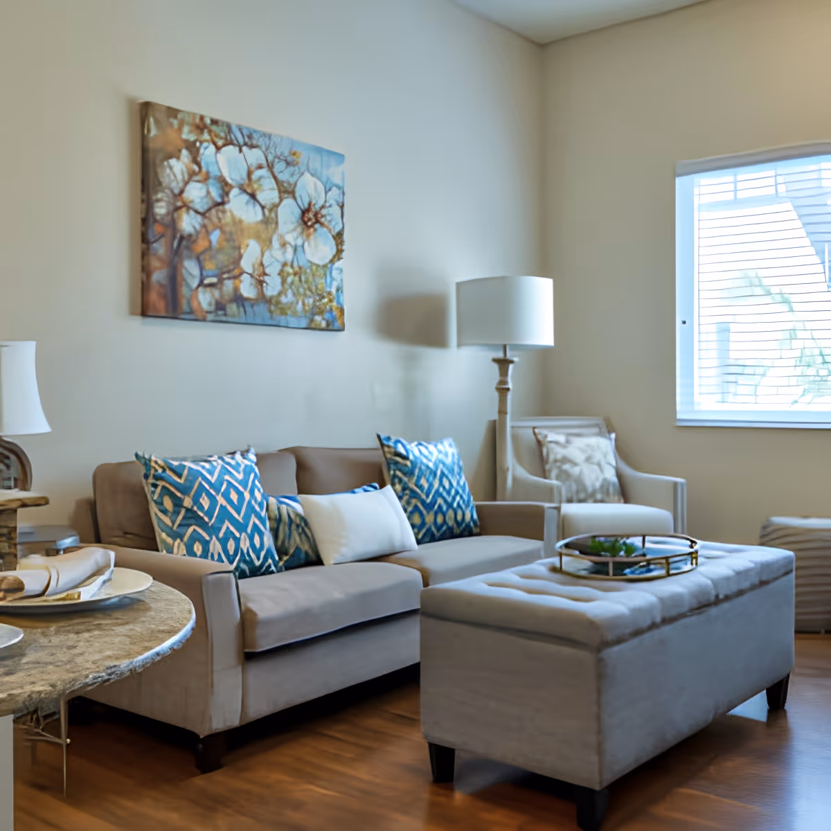 A cozy living room with a beige sofa adorned with blue and white patterned pillows, a matching armchair, a tufted ottoman with a decorative tray, a floor lamp, and a window with blinds letting in natural light. A floral painting hangs on the wall above the sofa.