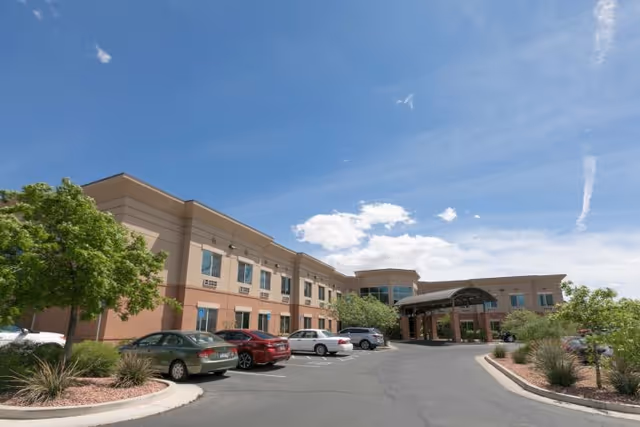 Exterior view of a two-story senior living facility building under a blue sky with some clouds. The building has beige and light brown walls with multiple windows. There is a covered entrance with a curved roof supported by columns. Several cars are parked in the parking lot in front of the building, and there are landscaped areas with trees and shrubs along the driveway.