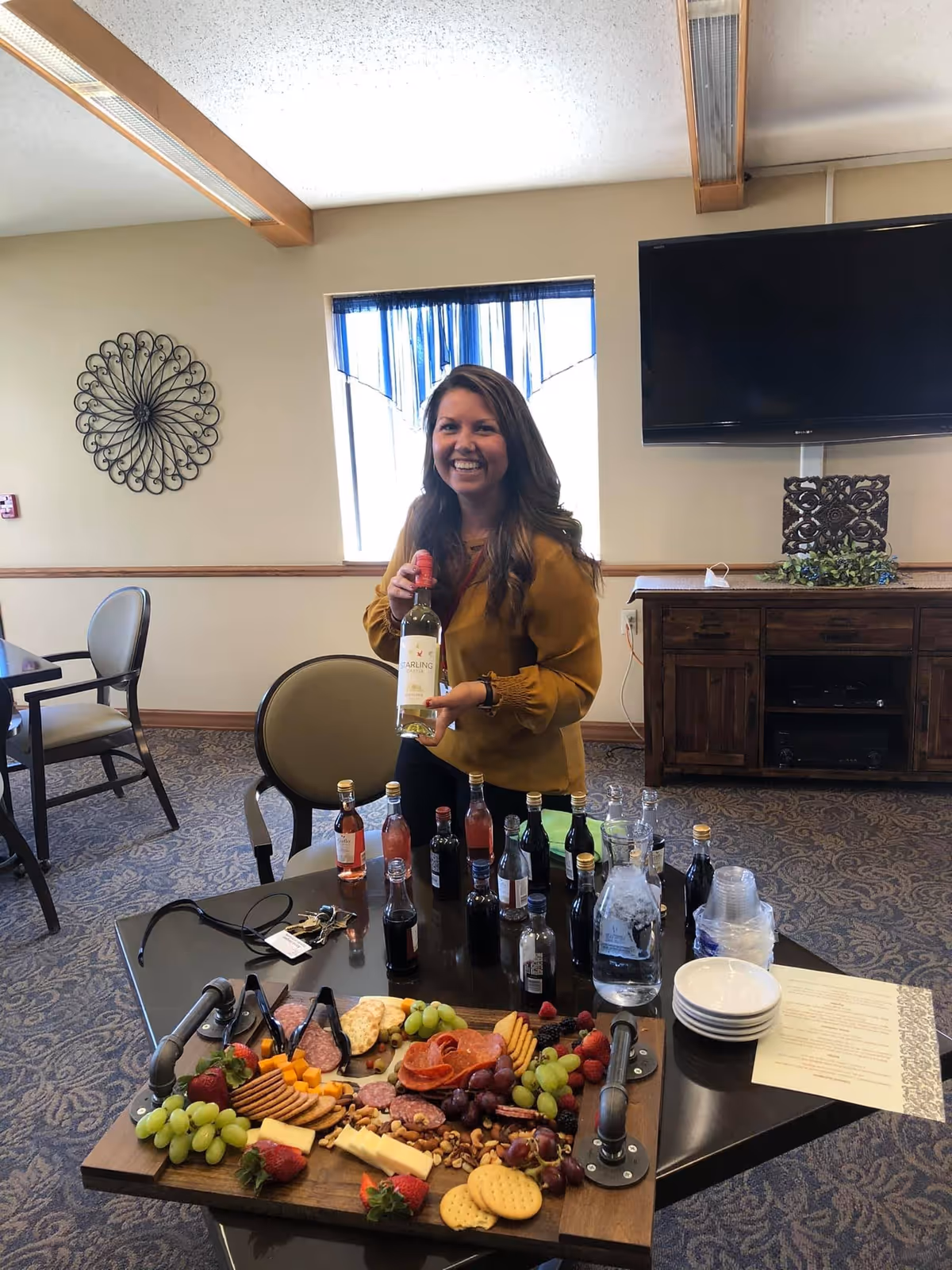 A smiling woman in a mustard yellow blouse stands behind a table with a charcuterie board featuring grapes, strawberries, cheese, crackers, and nuts. The table also has several small bottles of beverages, a pitcher of water with ice, stacked plastic cups, and a stack of small plates. The room has carpeted flooring, a wall-mounted TV, a window with blue curtains, and decorative wall art.