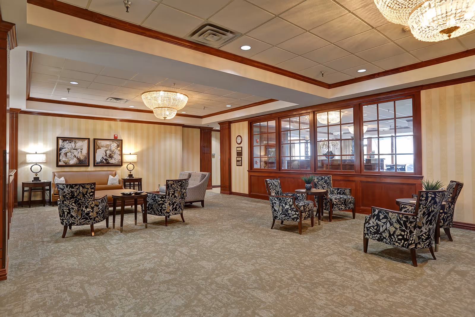 A spacious, well-lit common area with beige patterned carpet and cream-colored walls with wooden trim. The room features multiple seating arrangements with floral upholstered chairs and small wooden tables. There are two framed abstract paintings on the far wall above a beige sofa, flanked by two side tables with lamps. A large chandelier hangs from the ceiling, and a wooden partition with glass windows separates this space from another room.