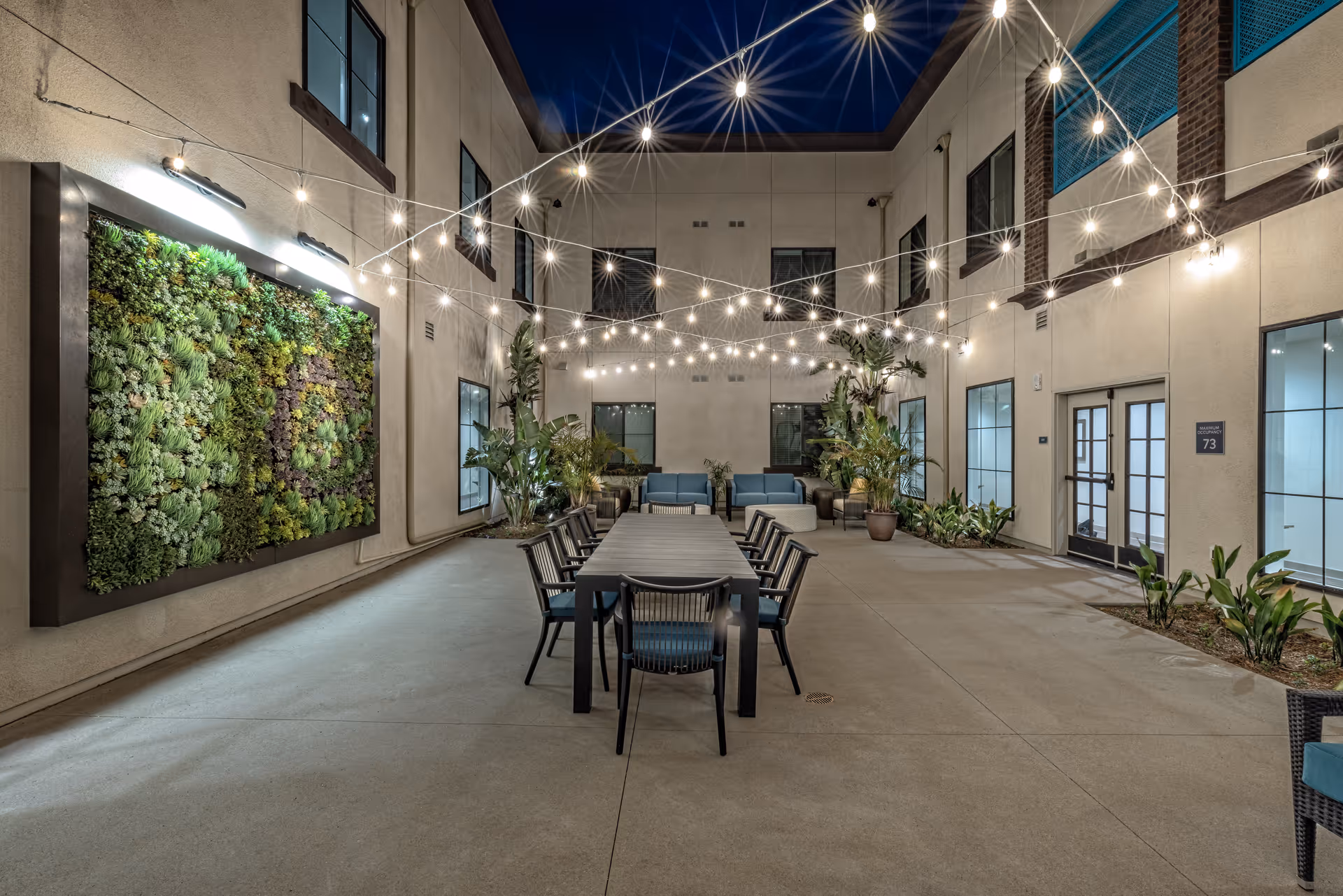 Outdoor courtyard area at night with string lights overhead, a long table with chairs in the center, potted plants along the walls, and a large vertical garden on one wall.