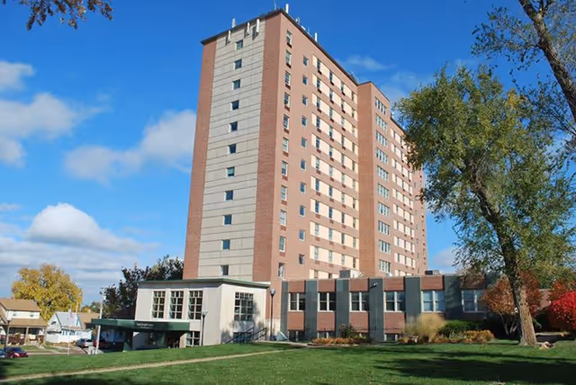 Exterior view of a multi-story brick assisted living tower with a lawn and trees under a blue sky.