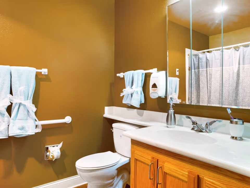 A bathroom with a toilet, sink and mirror, wooden vanity, towel bars holding blue towels, and a shower curtain.