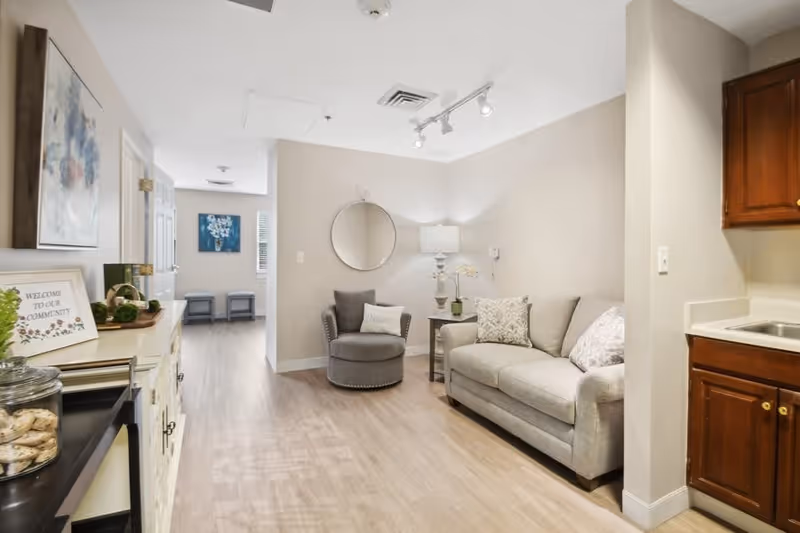 A cozy interior living area in a senior living facility with a gray sofa adorned with patterned pillows, a round gray armchair with a pillow, a side table with a lamp and decorative flowers, and a round mirror on the wall. The room has light wood flooring and neutral-colored walls. To the right, there is a small kitchenette with wooden cabinets and a sink. On the left side, a cabinet with a welcoming sign and some decorative items is visible, and a hallway leads to another room with two stools and a blue floral painting on the wall.