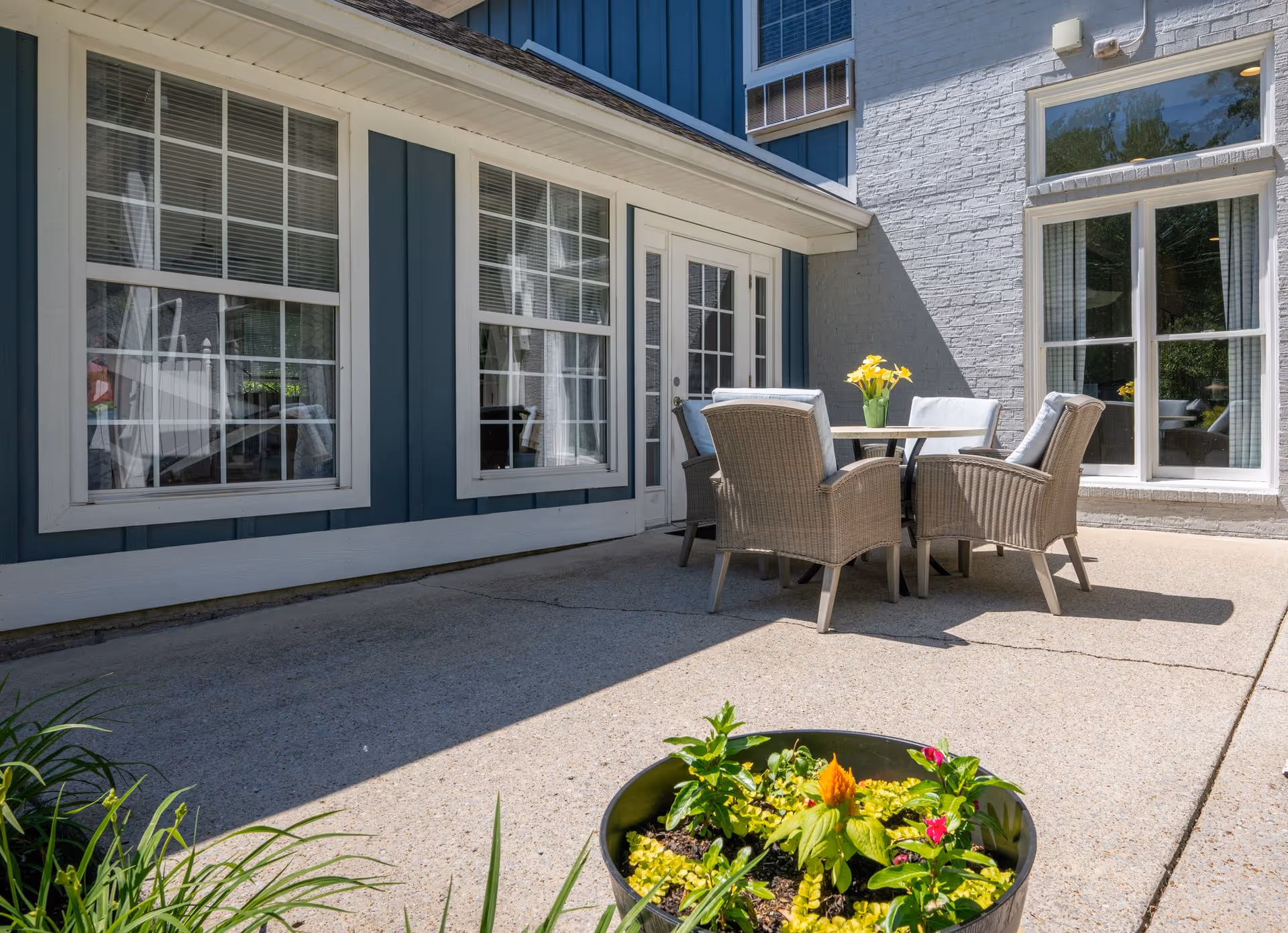 Outdoor patio area with a round table and four cushioned wicker chairs. A vase with yellow flowers is on the table. The patio is adjacent to a building with blue and white siding and multiple windows. A planter with green and flowering plants is in the foreground.