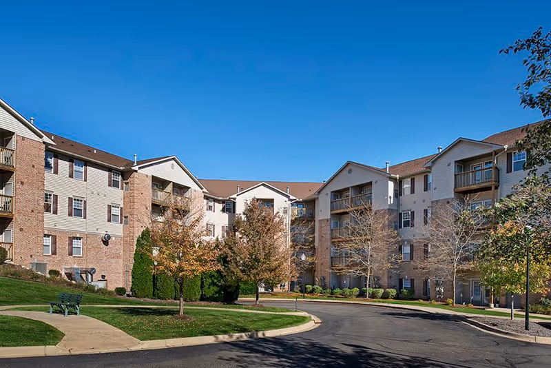 Exterior view of a multi-story senior living facility with beige and brick facade, balconies, trees with autumn foliage, a curved driveway, and a bench on a grassy area under a clear blue sky.