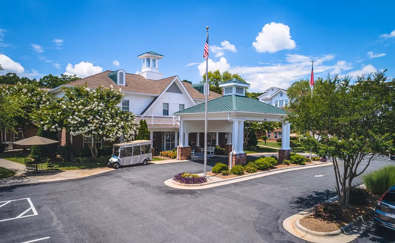 Exterior view of a senior living facility with a covered entrance supported by white columns and a green roof. There is an American flag on a flagpole in front of the entrance, a golf cart parked nearby, and trees and shrubs surrounding the building under a partly cloudy blue sky.