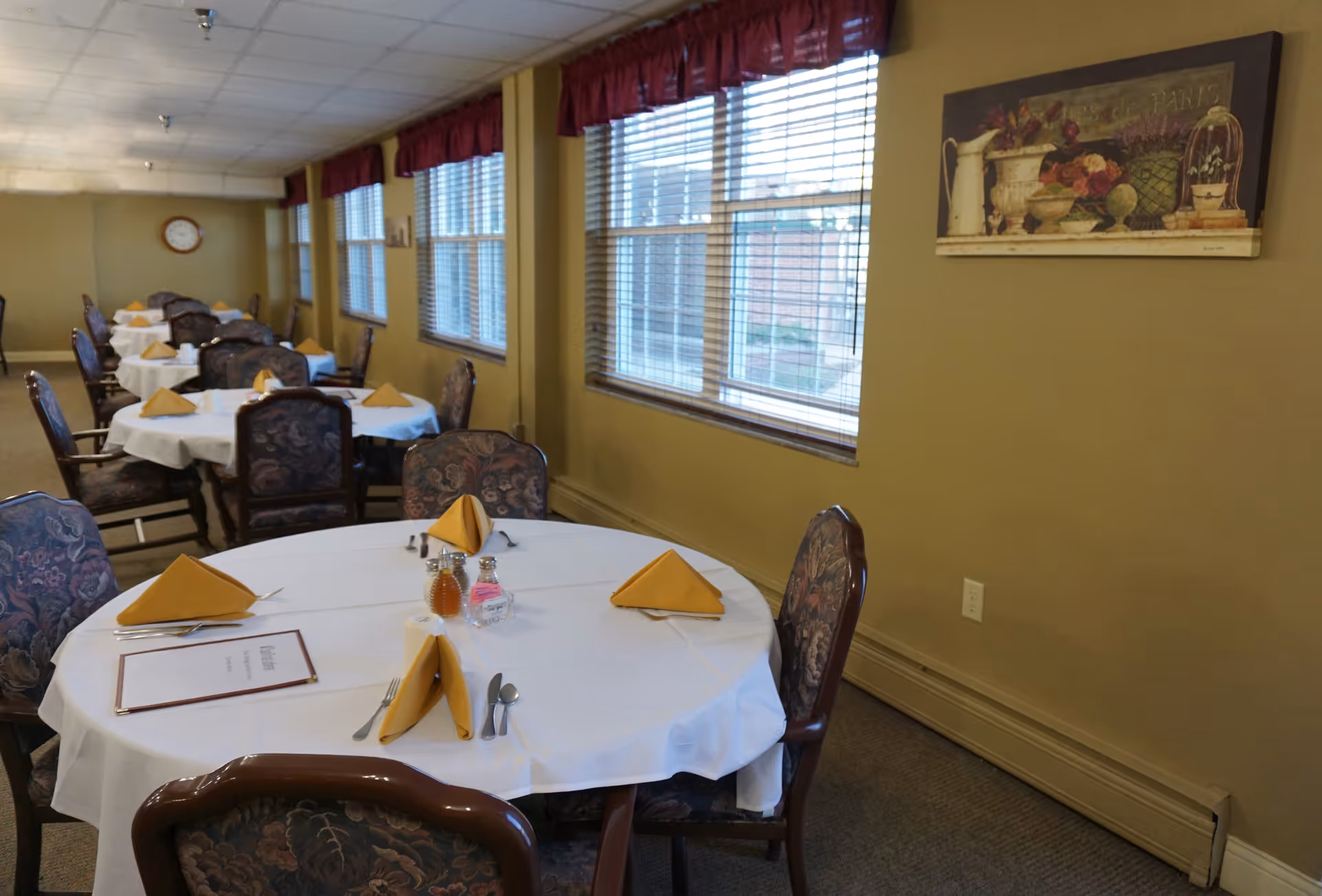 Dining room with round tables covered in white tablecloths, each set with yellow folded napkins, silverware, and condiments. The room has multiple windows with blinds and red valances, floral upholstered chairs, and a painting on the wall.
