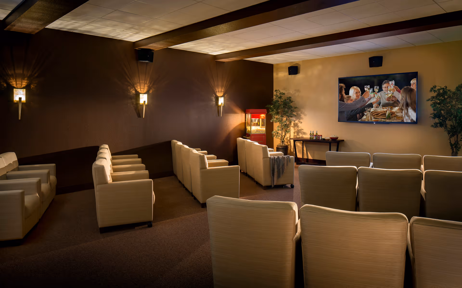 A cozy media room with several rows of beige armchairs facing a large flat-screen TV mounted on a beige wall. The TV displays a scene of people toasting with drinks. The room has warm lighting with wall sconces casting light patterns on a dark brown accent wall. There is a red popcorn machine and a small table with snacks and drinks near the TV, along with two potted plants on either side.
