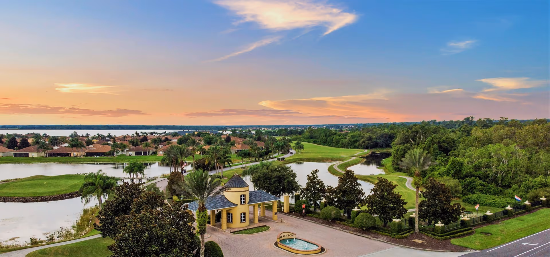 Aerial view of a scenic residential community with a small yellow gatehouse, surrounded by lush greenery, palm trees, water features, and a golf course under a colorful sunset sky.