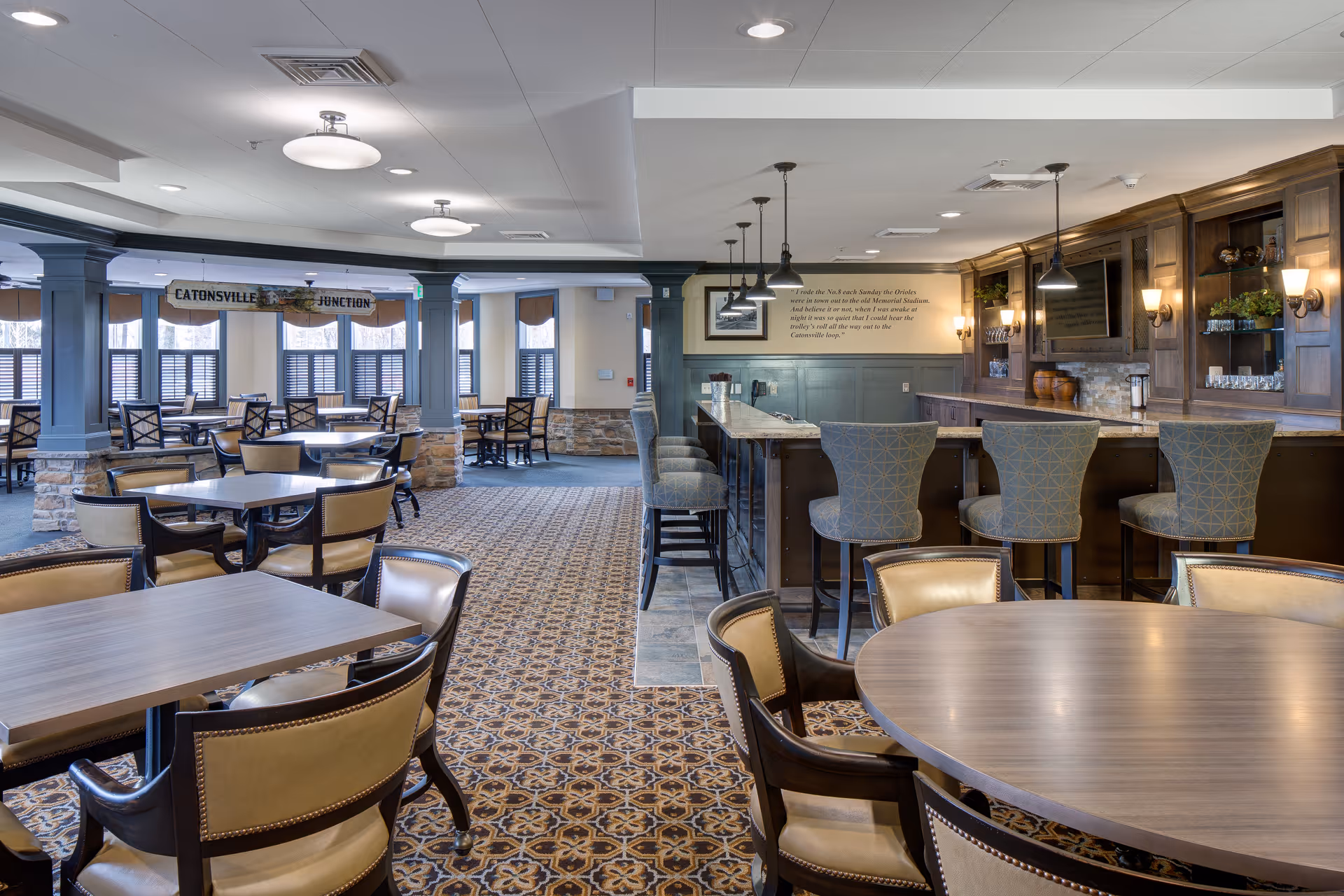 A spacious dining area in a senior living facility featuring multiple tables and chairs arranged on a patterned carpet. To the right, there is a bar counter with high chairs and pendant lighting above. The walls have wooden paneling and decorative sconces, with a television mounted behind the bar. Large windows with shutters allow natural light into the room.