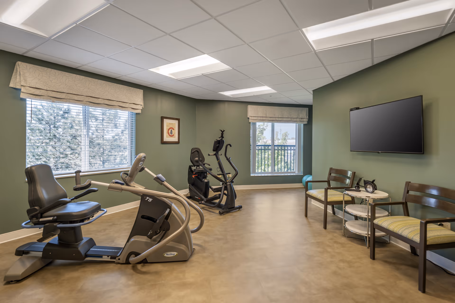 A small exercise room with two stationary exercise bikes positioned near two large windows with beige valances. The walls are painted green and the floor has a light brown finish. On the right side, there are two wooden chairs with green cushions and a small round table between them, holding a clock and a phone. A flat-screen TV is mounted on the wall above the chairs.