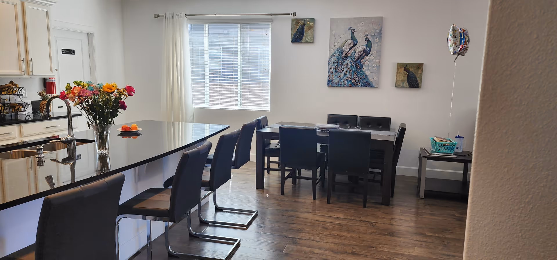 Open kitchen and dining area with a long black granite island and barstools, a dining table and chairs beneath peacock artwork, and a window.