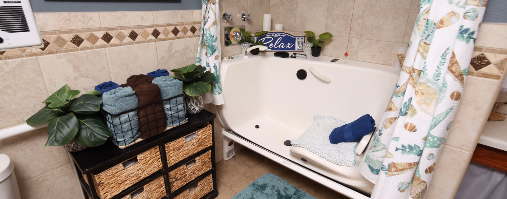 A bathroom with a white walk-in bathtub featuring a seat and grab bar. The tub area is decorated with a 'Relax' sign, small potted plants, and rolled towels. There is a black storage unit with woven drawers holding more rolled towels and plants. The walls are tiled with beige tiles and a decorative border, and a patterned shower curtain is partially drawn to the side.